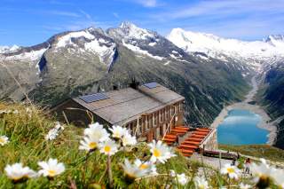 Hütte mit Blumenwiese und Blick auf Stausee und schneebedeckte Gipfel