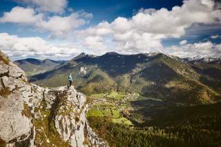 Eine Frau steht auf dem Leonhardstein und blickt ins Bergpanorama und auf das Bergsteigerdorf Kreuth im Tal.