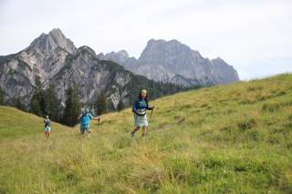 Recht zügig und mit Stöcken wandern drei Personen über eine Bergwiese. Im Hintergrund hohe Gipfel.
