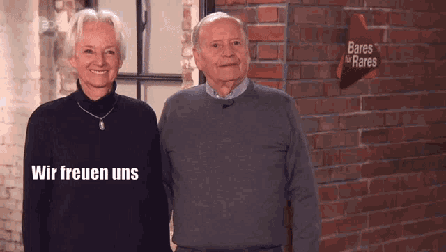 Ein älteres Paar steht lächelnd vor einer Backsteinwand mit der Aufschrift „Bares für Rares“. Neben ihnen erscheint der Schriftzug „Wir freuen uns“.