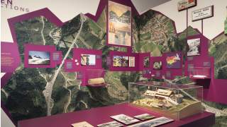 Ausstellungswand mit lila Elementen, historischen Fotos, Karten, einem Poster, Infotafeln und einem Modell von Berghütten auf einer Luftaufnahme einer Gebirgslandschaft.