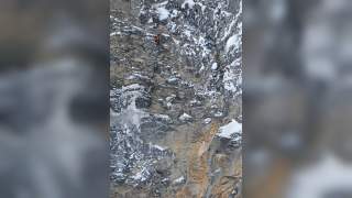 Ein Kletterer mit orangefarbener Jacke erklimmt eine steile, schneebedeckte Felswand.
