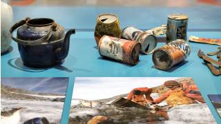 Alte, verrostete Konservendosen, ein blauer Teekessel und Besteck liegen auf einer blauen Ausstellungsfläche; vorne Fotos von Menschen bei einer Aufräumaktion in einer Berglandschaft, wo u. a. genau jener Kessel geborgen wurde.