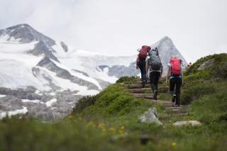 Eine Wandergruppe unterwegs im Hochgebirge – im Vordergrund grüne Wiesen, im Hintergrund vergletscherte Berge.