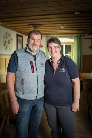 Ein Mann und eine Frau stehen lächelnd nebeneinander in einem gemütlichen, holzvertäfelten Gastraum.