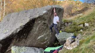 Ein Mann klettert an einem großen Felsblock im Freien und hält sich mit einer Hand oben fest; am Boden liegen Bouldermatten und Ausrüstung.