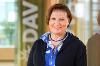 Porträt einer lächelnden Frau mit kurzen braunen Haaren, die eine blaue Bluse, eine schwarze Strickjacke und eine blaue Perlenkette trägt.