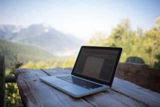 Im Vordergrund ist ein Laptop auf einem Holztisch zu sehen, im Hintergrund unscharf Berge.