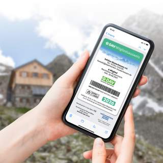 Hand hält Smartphone mit digitalem Mitgliedsausweis des Deutschen Alpenvereins, im Hintergrund unscharfe Berglandschaft mit Hütte.