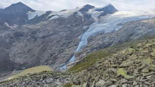 Steiniger Berghang im Vordergrund, dahinter ein Gletscher, der von felsigen Gipfeln ins Tal fließt, unten ein kleiner, milchig-grüner Gletschersee.
