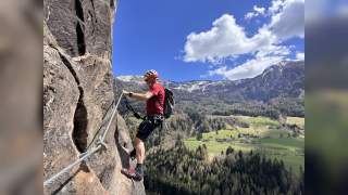 Ein Klettersteiggeher mit Helm und Ausrüstung steht an einer Felswand, im Hintergrund Berge, Wälder und ein Tal; über dem Mann ein Metallzeichen mit der Aufschrift „Ratschings Racines“ und einem Steinbock.