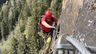 Eine Person mit rotem Helm und rotem T-Shirt klettert gesichert an einem Stahlseil eine steile Felswand hinauf, im Hintergrund dichter Nadelwald.
