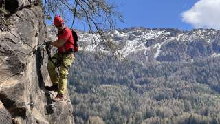 Person mit Klettersteigausrüstung und rotem Helm klettert an einer Felswand, im Hintergrund schneebedeckte Berge und bewaldete Hänge unter blauem Himmel.