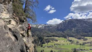 Ein Klettersteiggeher mit Helm und Ausrüstung steht an einer steilen Felswand, im Hintergrund grüne Täler, Wälder und schneebedeckte Berge unter blauem Himmel mit Wolken.