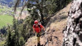 Person mit Klettersteigausrüstung und Helm geht an einem Drahtseil auf einem steilen, bewaldeten Berghang entlang, im Hintergrund grüne Felder und ein Tal.
