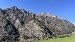 Grüne Wiese im Vordergrund, dahinter ein felsiger, teils bewaldeter Berghang unter klarem, blauem Himmel.