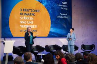 Die Moderatorin Salwa Houmsi und eine Referentin stehen auf der Bühne, im Hintergrund ist das Titelbild der Veranstaltung mit dem Motto 