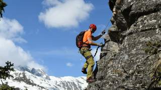 Ein Klettersteiggeher mit Helm und Ausrüstung erklimmt eine Felswand, im Hintergrund schneebedeckte Berge und blauer Himmel.
