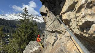 Eine Person mit Klettersteigausrüstung und rotem Helm klettert an einer Felswand entlang eines gesicherten Klettersteigs in den Bergen, im Hintergrund verschneite Gipfel und Nadelwald.