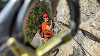Ein Kletterer mit rotem Helm und orangefarbenem Shirt ist an einer Felswand gesichert; im Vordergrund sind Karabinerhaken unscharf zu sehen, im Hintergrund Berge und Bäume.