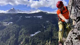 Klettersteiggeher mit Helm und Ausrüstung erklimmt eine Felswand, im Hintergrund verschneite Berge und ein dichter Nadelwald.