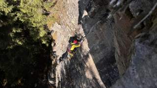 Eine Person mit Kletterausrüstung und rotem Helm klettert an einer steilen Felswand, im Hintergrund sind Bäume zu sehen.