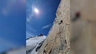 Bergsteiger klettert an einer steilen Felswand, im Hintergrund strahlende Sonne und schneebedeckter Gipfel unter blauem Himmel.