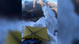 Gelbes Zelt steht auf schneebedecktem, steilem Berghang mit eisigen Felsen; im Hintergrund Berge und ein kleiner blauer See im Tal.