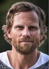 Porträt eines lächelnden Mannes mit kurzem braunem Haar und Vollbart vor unscharf grünem Hintergrund.