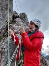 Eine Person in roter Outdoorjacke und Kletterhelm steht an einer Felswand und hält mehrere Kletterseile in den Händen, während ein Seil nach oben verläuft; im Hintergrund sind graue Wolken und Berglandschaft zu sehen.