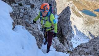 Bergsteigerin mit Helm und Kletterausrüstung steigt einen verschneiten, felsigen Hang hinauf; im Hintergrund ein Tal mit Bergsee.