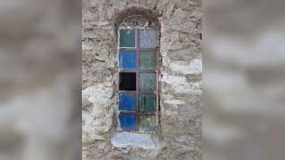 Schmales, hochrechteckiges Fenster mit bunten, teils zerbrochenen Glasscheiben in einer rauen, grauen Steinmauer.