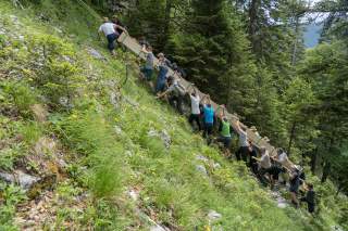Männer hieven den Längsbalken des neuen Gipfelkreuzes über einen steilen Berghang.