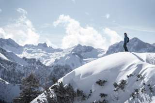 Person mit Wanderstöcken steht auf einem verschneiten Hügel vor einer Bergkulisse.