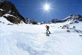 Person mit Skiausrüstung wandert bei sonnigem Himmel und schneebedeckten Bergen im Hintergrund einen Hang hinauf.