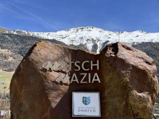 Großer Fels mit der Aufschrift „Matsch Mazia“ und einem Schild „Bergsteiger Dörfer“ vor schneebedecktem Berg und blauem Himmel.