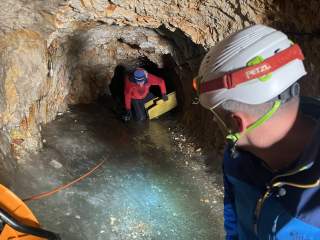 Zwei Personen mit Helmen erkunden einen engen, felsigen Tunnel, wobei eine Person kniend eine gelbe Tafel hält und die andere mit Stirnlampe den Tunnel betrachtet. Der Boden ist teils mit Eis bedeckt.