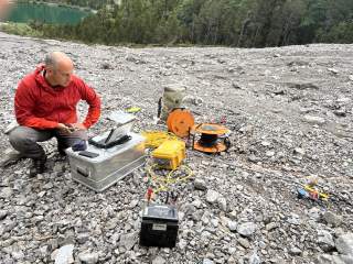 Ein Mann in roter Jacke kniet auf einem felsigen Boden und bedient technische Messgeräte mit Kabeln und einem Laptop, im Hintergrund sind Wald und See zu sehen.