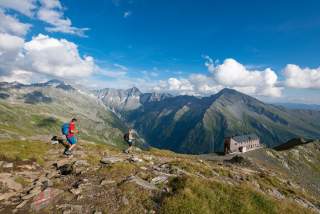 Zwei Wanderer auf einem Bergpfad in einer alpinen Landschaft, im Hintergrund Berge und eine Berghütte unter blauem Himmel mit wenigen Wolken.