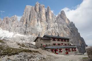 Steinernes Berghaus mit rot-weißen Fensterläden steht vor steilen, felsigen Gipfeln und blauem Himmel.