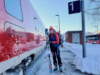 Frau in Winterkleidung mit Skiern und Skischuhen steht an einem schneebedeckten Bahnsteig neben einem roten Zug. Ein Schild mit der Nummer 1 ist rechts im Bild zu sehen.
