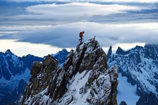 Bergsteiger mit rotem Rucksack erklimmt schneebedeckten, felsigen Grat, umgeben von hohen, verschneiten Bergen und dramatischem Wolkenhimmel.