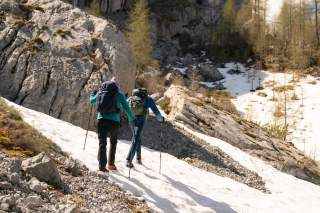 Zwei Menschen wandern mit Rucksäcken und Wanderstöcken einen verschneiten Hang in den Bergen hinauf.