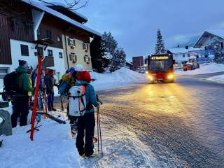 Menschen mit Skiausrüstung warten an einer Schnee bedeckten Straße auf einen ankommenden Bus mit der Anzeige „47 Sonthofen“. Hintergrund ist ein winterliches Dorf mit beleuchteten Gebäuden.