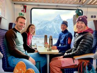 Vier lächelnde Personen in sportlicher Kleidung sitzen an einem Tisch im Zug, im Fensterrahmen ist eine verschneite Berglandschaft zu sehen.
