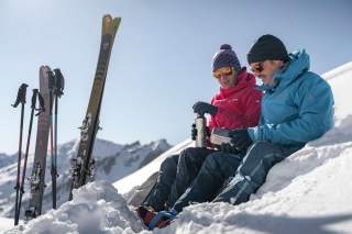 Zwei Personen in Winterkleidung sitzen im Schnee und machen eine Pause, während ihre Skier und Skistöcke neben ihnen im Schnee stecken.