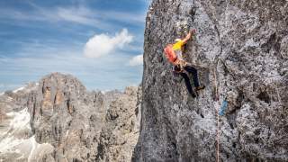 Ein Kletterer mit Helm und Seil klettert an einer steilen Felswand in den Bergen, im Hintergrund sind weitere felsige Gipfel und blauer Himmel zu sehen.