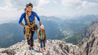 Zwei Frauen mit Kletterausrüstung und Helmen stehen auf einem felsigen Berggipfel, im Hintergrund weite Berglandschaft und Tal.
