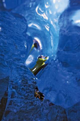 Blick durch blaues Eis auf eine Person in grüner Jacke mit Steigeisen, die in einer Eishöhle klettert.