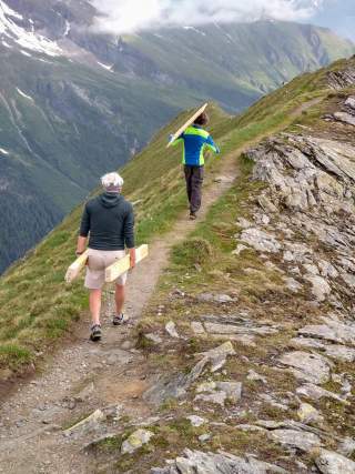 Ein Mann und eine Frau laufen auf einem Bergpfad, beladen mit Holzteilen.